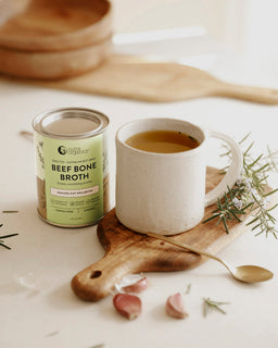 A mug of broth sits next to a tin of Nutra Organic's Beef Bone Broth garden herb