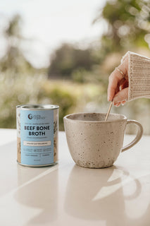 A mug of broth being stirred sits next to a tin of Nutra Organic's Beef Bone Broth hearty original